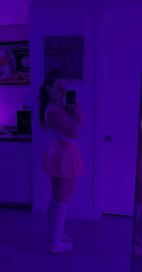 Woman in white top and skirt, thigh-high socks, taking mirror selfie in purple-lit room.