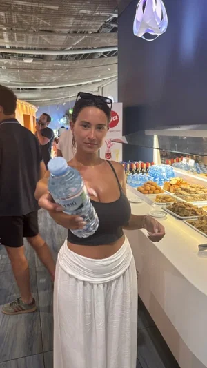 Woman in black crop top and white skirt holding water bottle at food stall.