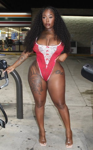Woman with curly hair in red and white lace bodysuit, standing outdoors near gas pump.