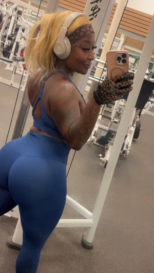 Woman in blue workout gear takes mirror selfie at gym, wearing headphones and leopard print gloves.