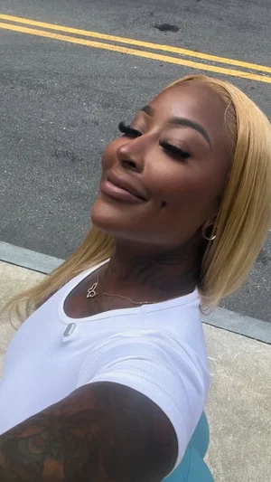 Woman with blonde hair and tattoos wears white top, takes outdoor selfie on street.