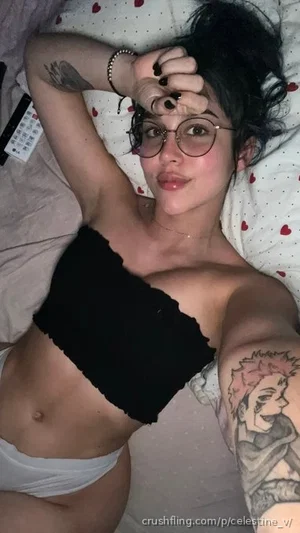 Woman with dark hair and glasses lying in bed, wearing black crop top and white underwear, tattoos