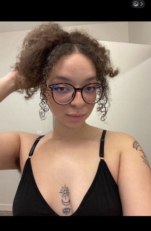 Woman with curly hair and glasses wears black top, shows chest tattoo, indoors.