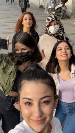 Five women in casual clothing pose for a group selfie on a city sidewalk.