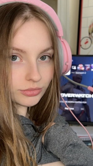 Woman with long brown hair wearing pink headphones and gray top, indoors near a monitor.
