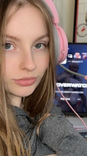 Woman with long brown hair wearing pink headphones and gray top, indoors near a screen.