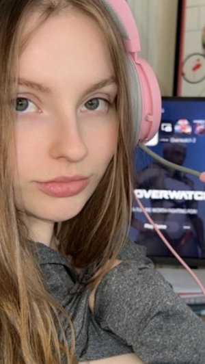 Woman with long hair wearing pink headphones and gray top, indoors near a monitor.