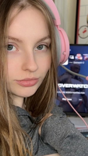 Woman with long brown hair wearing pink headphones and gray top, indoors near a monitor.