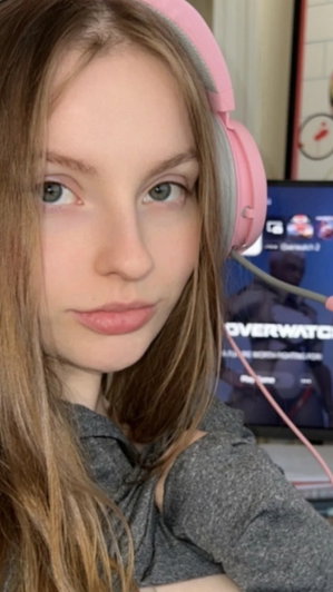 Woman with long brown hair wearing pink headphones and gray top, indoors near a computer screen.