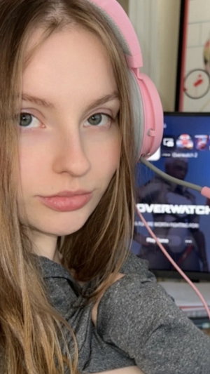 Woman with long brown hair wearing pink headphones and gray top, indoors near a monitor.