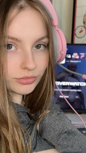 Woman with long brown hair wearing pink headphones and gray top, indoors near a screen.