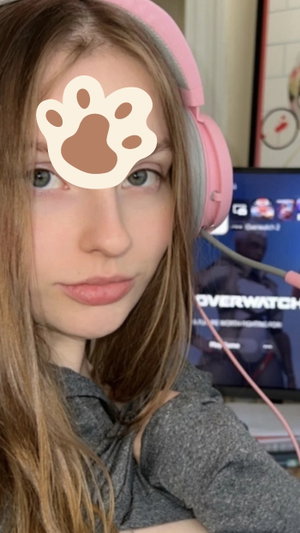 Woman with long hair wearing pink headphones and gray top, indoors near a monitor.