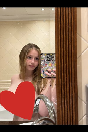 Woman with wet hair taking mirror selfie in bathroom, bare shoulders, red heart overlay.