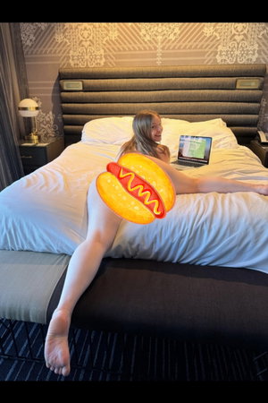 Woman with blonde hair lying on bed, wearing no visible clothing, with hot dog emoji covering lower
