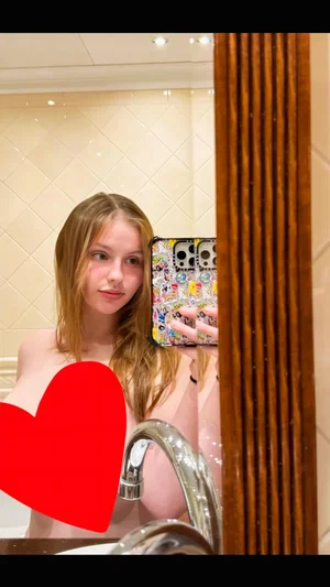 Woman with long hair taking mirror selfie in bathroom, bare shoulders, red heart overlay.