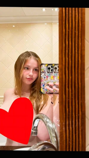 Woman with long blonde hair taking mirror selfie in bathroom, bare shoulders, red heart overlay.