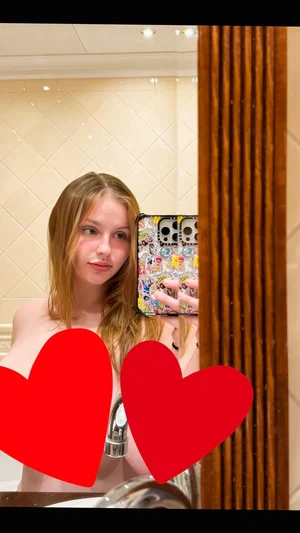 Woman with long blonde hair taking mirror selfie in bathroom, bare shoulders, red hearts covering