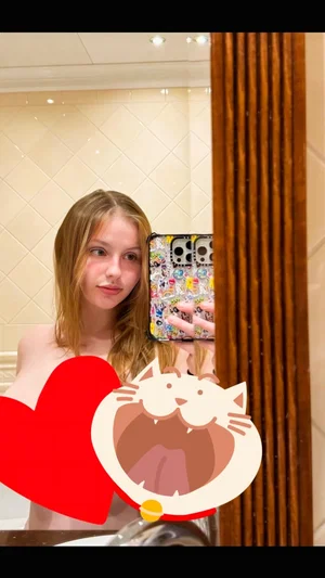 Woman with light hair in bathroom mirror selfie, topless, holding phone with stickers.