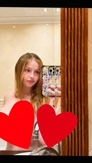 Woman with long blonde hair taking mirror selfie in bathroom, bare shoulders, red hearts covering