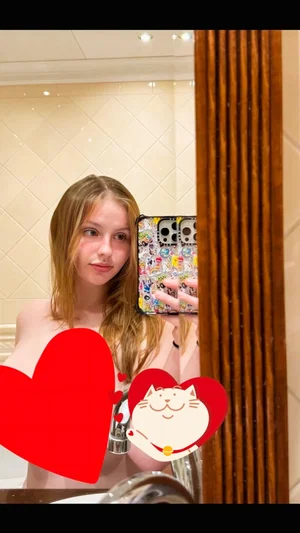 Woman with long hair taking mirror selfie in bathroom, wearing red heart and cat stickers.