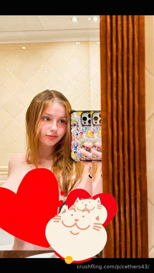 Woman with long hair taking mirror selfie in bathroom, wearing no top, red heart and cat sticker