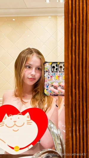 Woman with blonde hair taking mirror selfie in bathroom, wearing no top, holding phone with sticker