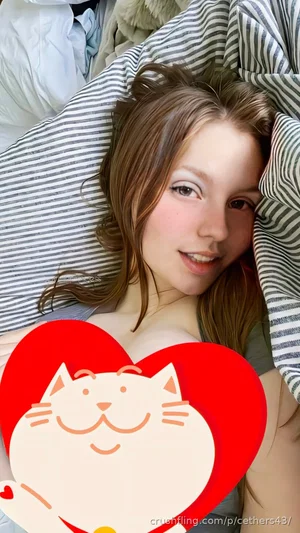 Woman with brown hair lying in bed, wearing gray top, smiling at camera.