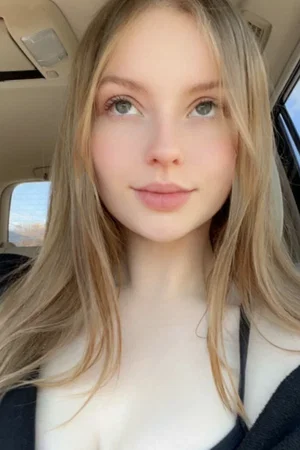 Blonde woman in car, wearing dark top, looking at camera.