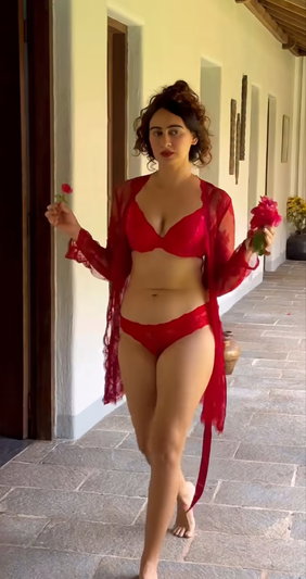 Woman in red lingerie and sheer robe holding flowers, walking on stone patio.