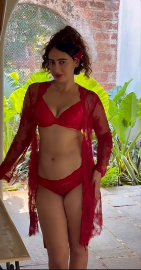 Woman in red lingerie and sheer robe standing outdoors near plants.