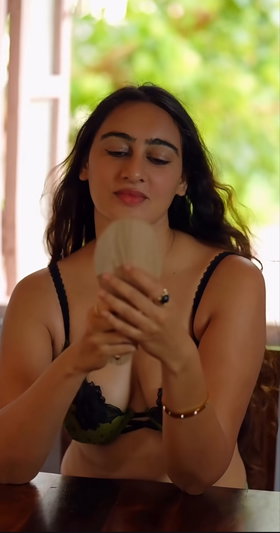 Woman in black lace bra holding mirror, sitting indoors near window with greenery outside.