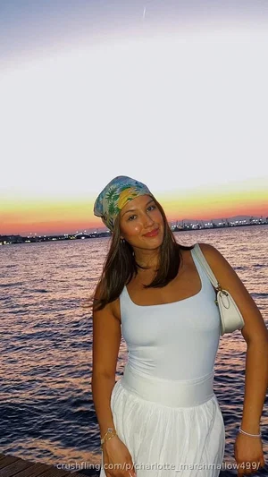 Woman in white top and skirt, wearing headscarf, standing by water at sunset.