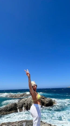Woman in yellow bikini top and white pants posing on rocky beach by ocean.