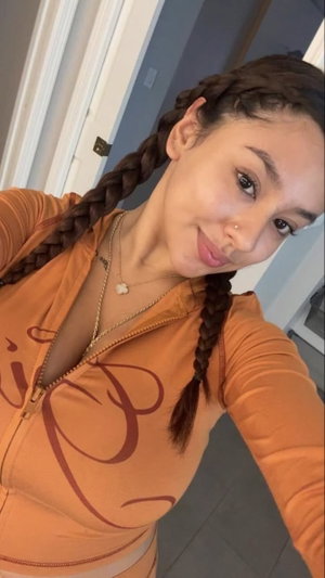 Woman with braided hair in orange zip-up top, indoors, smiling at camera.