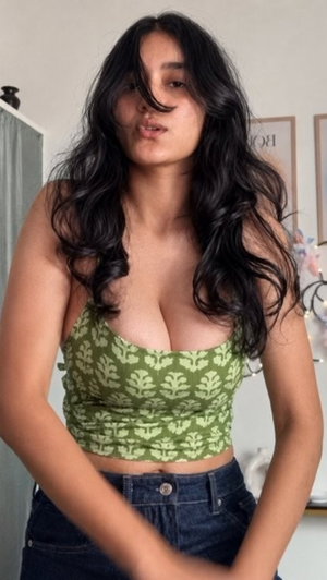 Woman with long dark hair wearing green patterned crop top and jeans indoors.