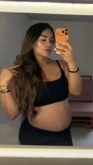 Woman with long brown hair in black crop top and pants, taking mirror selfie in indoor setting.