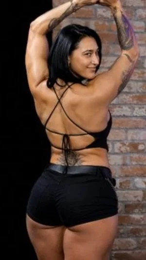 Woman with tattoos in black strappy top and shorts, posing in front of brick wall.