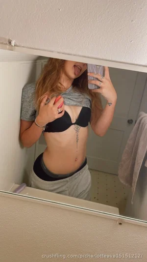 Woman with long hair in black bra and gray shirt, taking mirror selfie in bathroom.