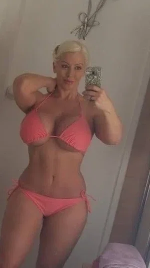 Blonde woman in pink bikini taking mirror selfie in indoor setting.