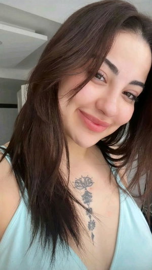 Woman with long dark hair and chest tattoo wearing light blue top indoors.