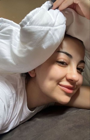 Woman with dark hair lying on bed, wearing white shirt, smiling at camera.