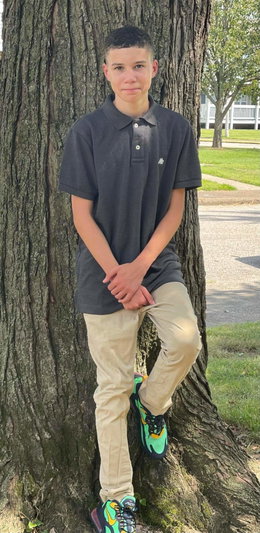 Male in dark polo shirt and khaki pants leaning on tree outdoors.