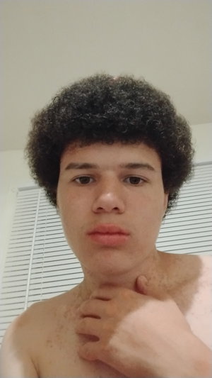Man with curly hair shirtless indoors touching neck.