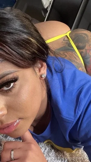 Woman with dark hair and makeup, wearing blue top and yellow straps, tattoo on shoulder, posing
