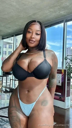 Woman with tattoos in black bra and light blue thong, posing indoors near large windows.