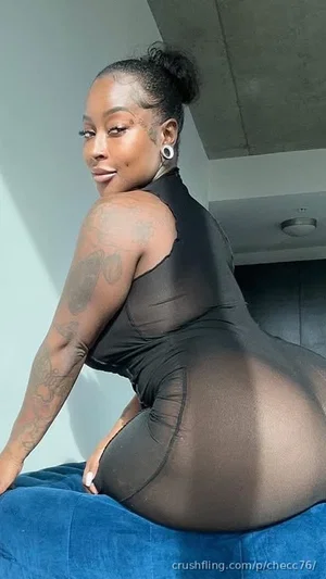 Woman in black sheer bodysuit posing on blue couch, tattoos on arm, indoor setting.