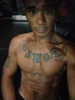 Shirtless tattooed man sitting indoors on dark furniture.