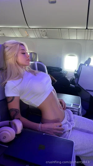 Blonde woman in white crop top and sweatpants sitting on airplane, hand on hip.