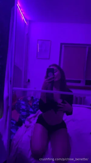 Woman in black lingerie posing on bed in purple-lit room, mirror selfie.