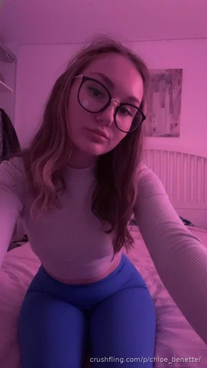 Woman with glasses in a white top and blue leggings sitting on a bed under pink lighting.